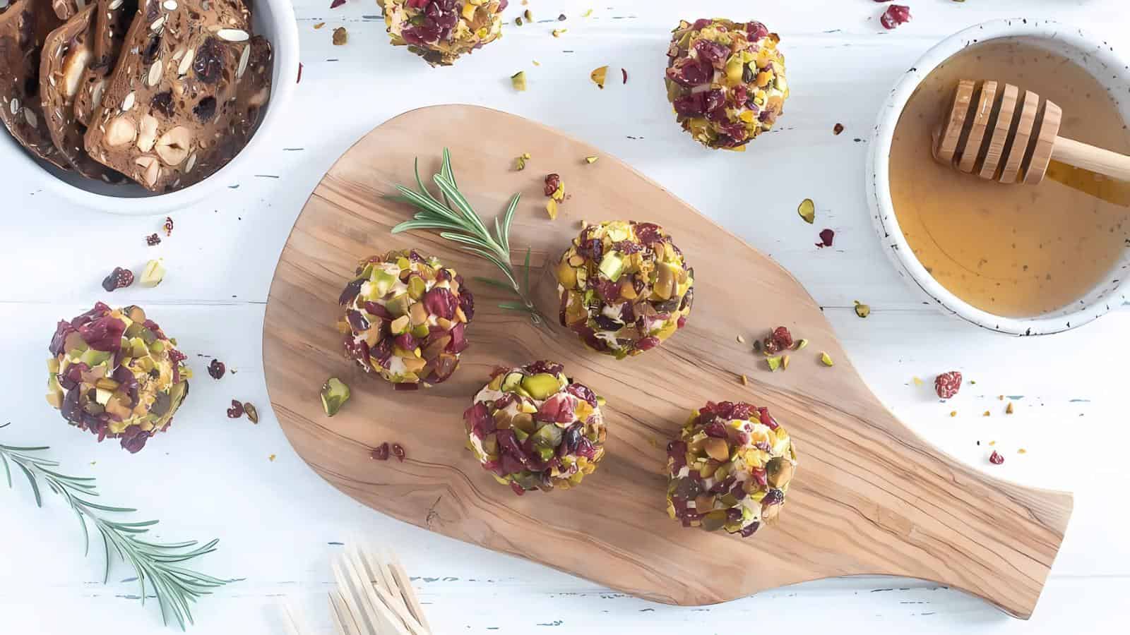 Cheese balls coated with nuts and dried fruit on a wooden board, with a bowl of honey, a sprig of rosemary, crispy crackers, and toothpicks nearby.