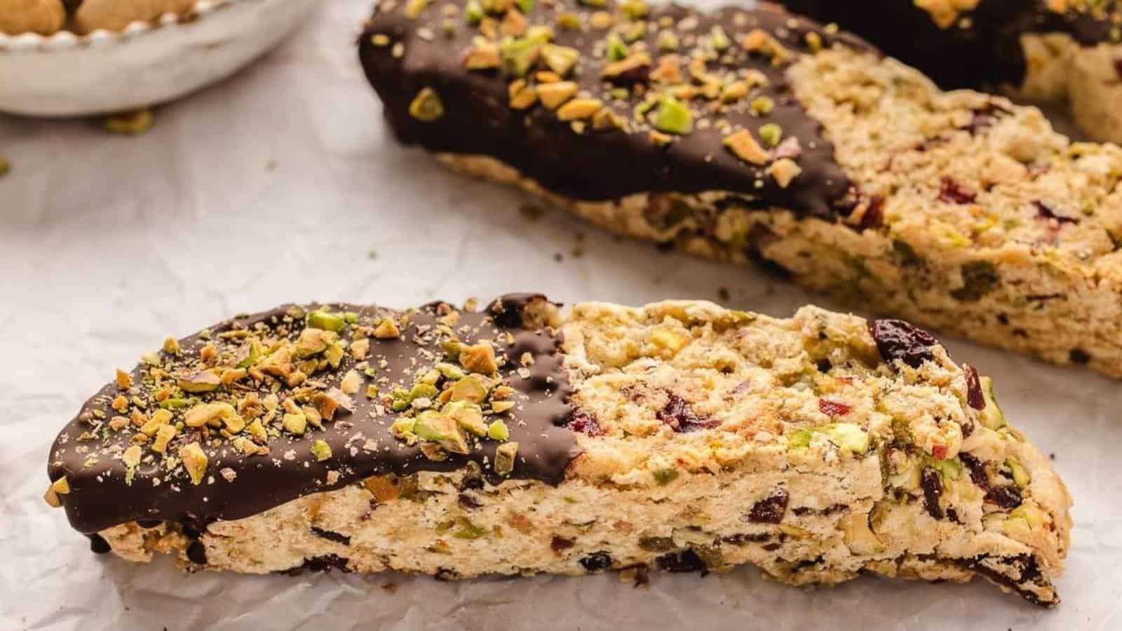 Close-up of biscotti partially dipped in chocolate, topped with crushed pistachios, placed on parchment paper.