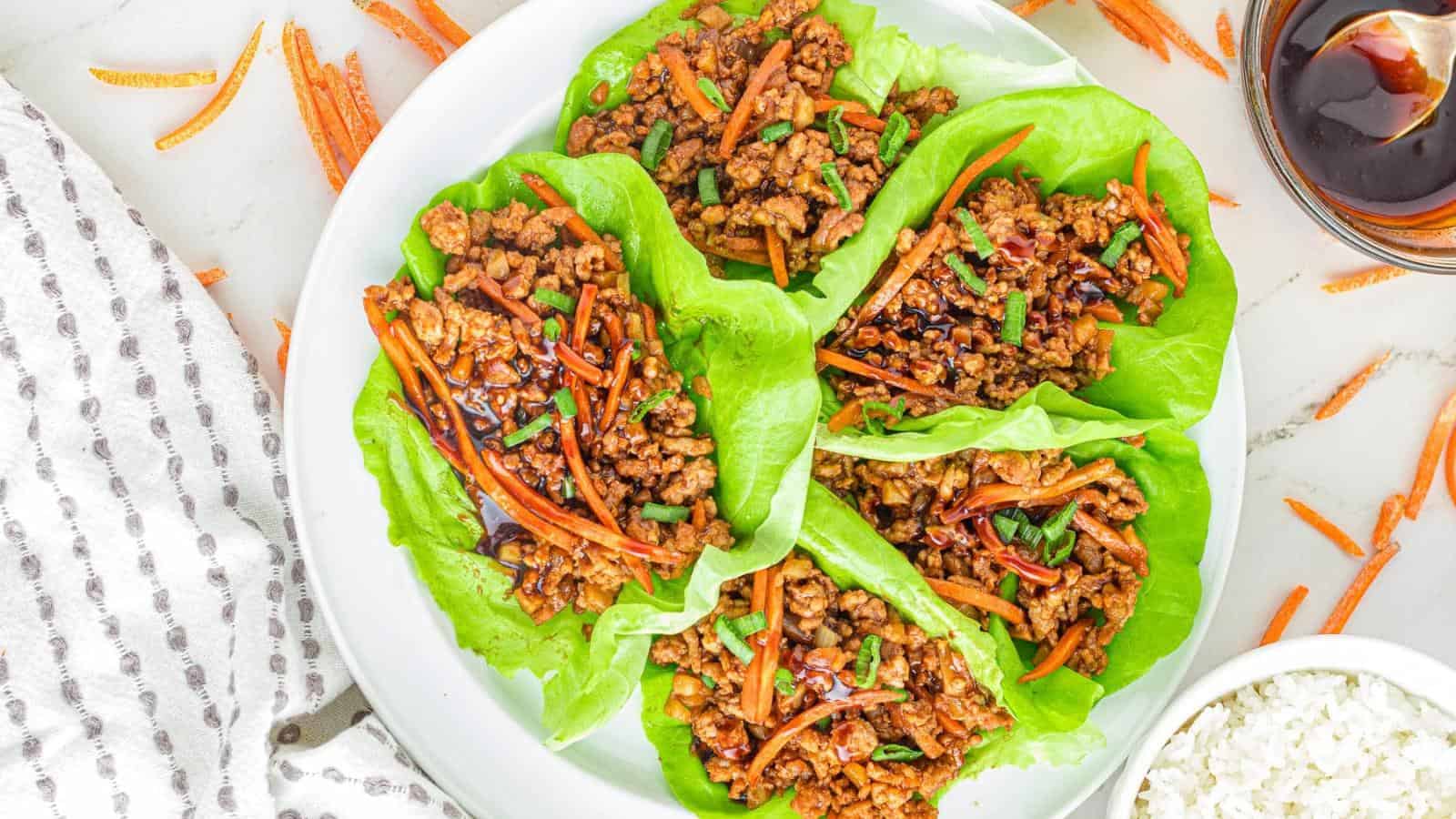 Four lettuce wraps filled with seasoned ground meat, shredded carrots, and garnished with herbs on a white plate, accompanied by a small bowl of sauce and a side of rice.