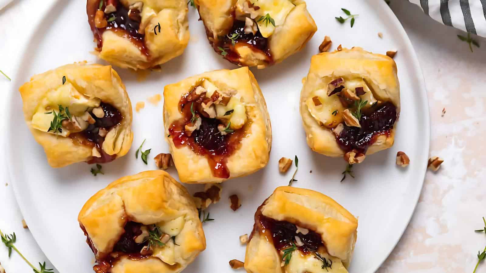 Seven puff pastry bites filled with brie cheese, jam, and chopped nuts on a white plate, garnished with herbs.