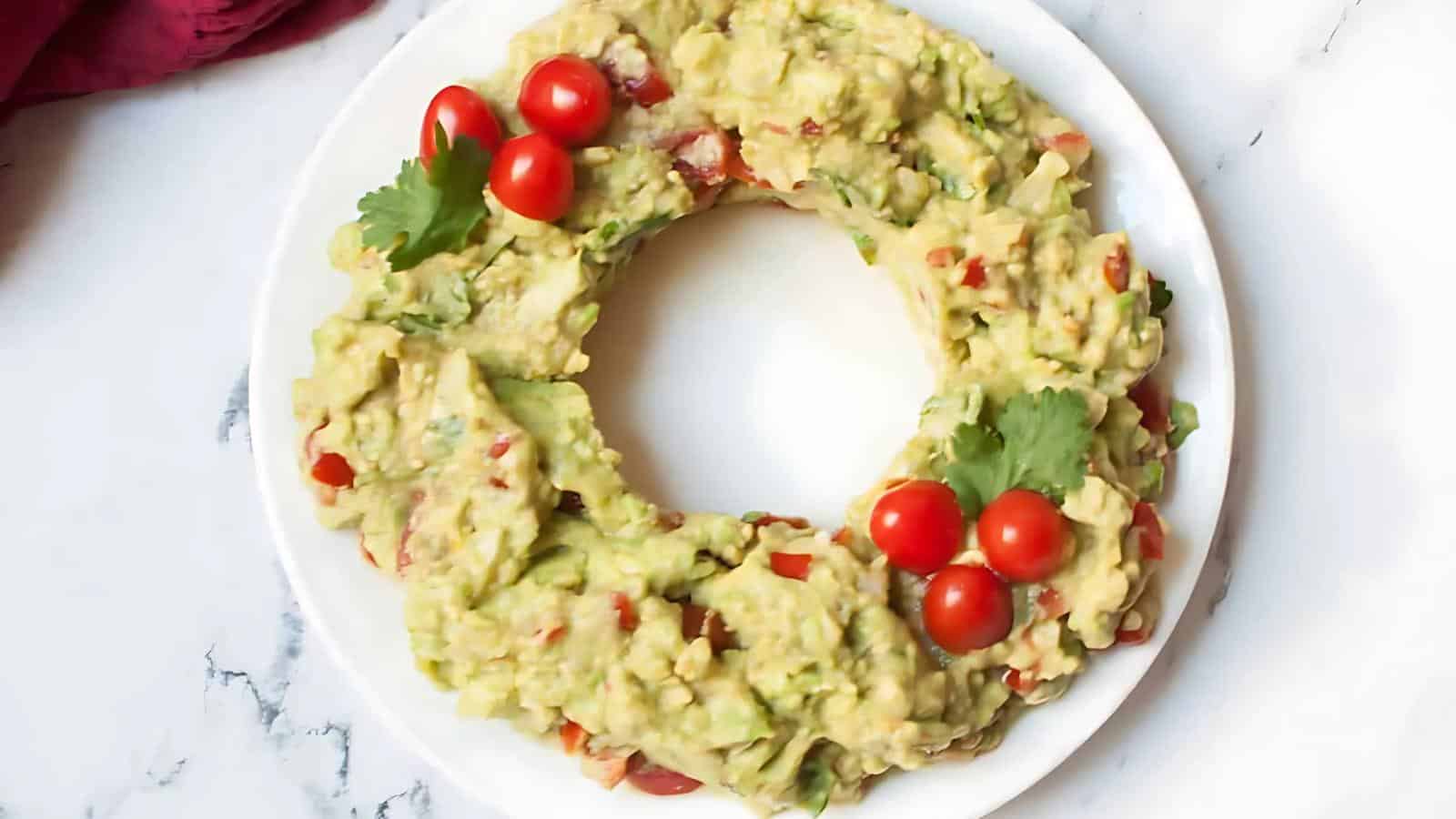 A plate of guacamole shaped like a wreath, garnished with cherry tomatoes and cilantro, on a marble surface.