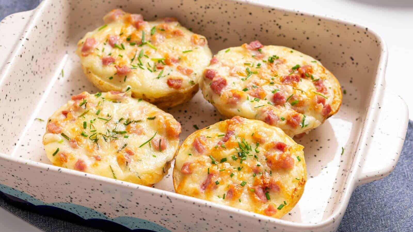 Four baked potato halves topped with melted cheese, diced ham, and herbs in a speckled baking dish.
