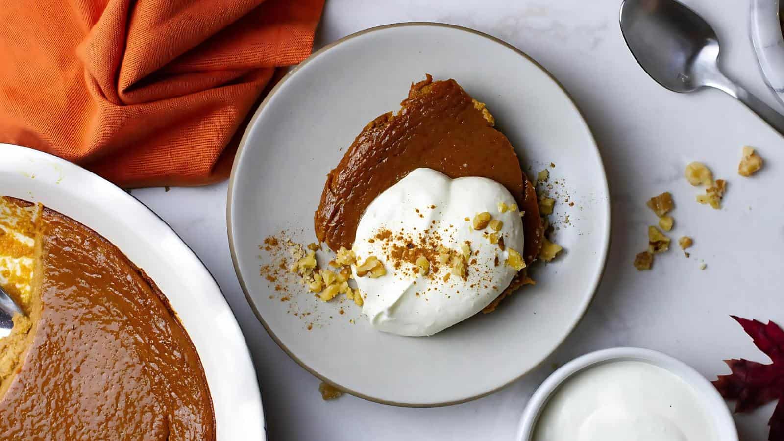 A slice of pumpkin pie topped with whipped cream and nuts on a plate. Orange cloth and a pie dish are nearby.