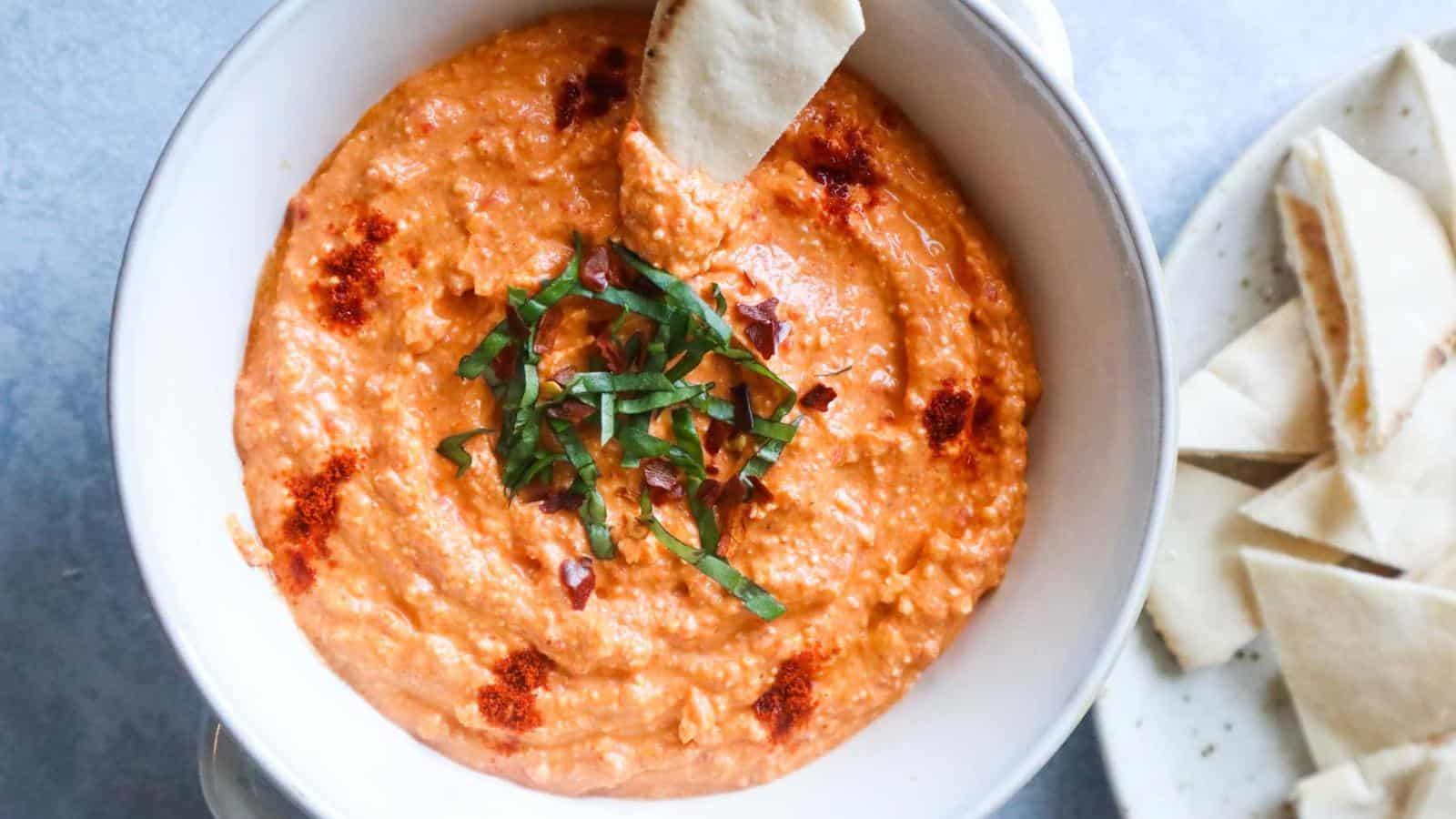 A bowl of red pepper hummus garnished with chopped herbs and spices, accompanied by pita bread slices.