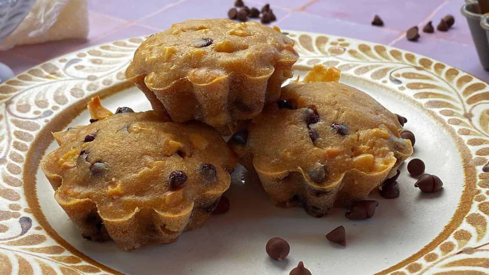 Three chocolate chip muffins stacked on a decorative plate, surrounded by scattered chocolate chips.