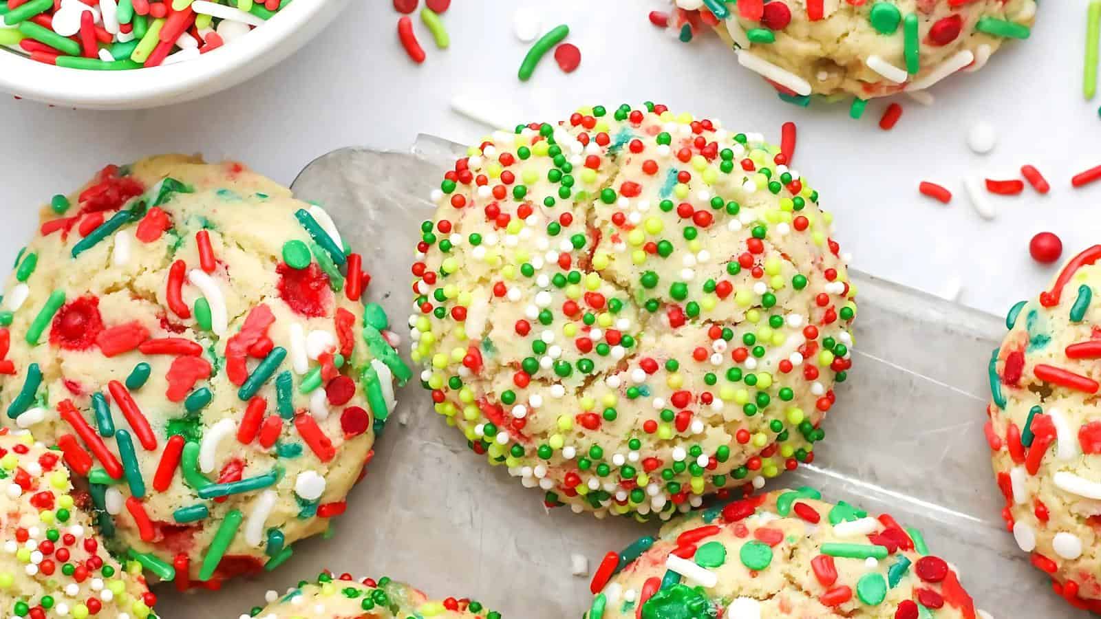 Colorful cookies with red, green, and white sprinkles on a tray, surrounded by more cookies and a bowl of sprinkles.