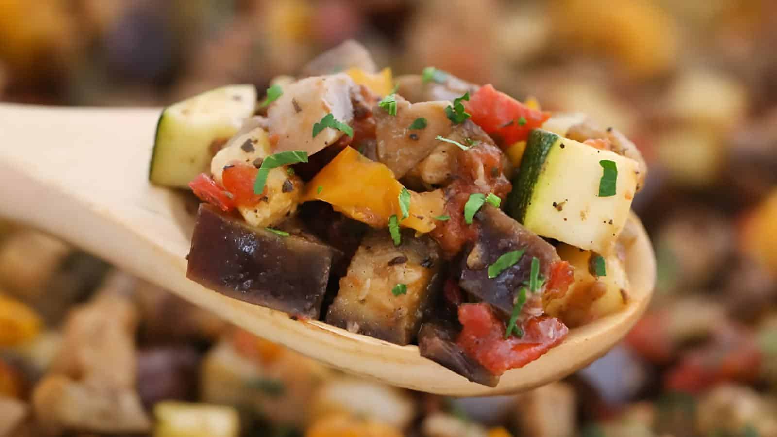 Close-up of a wooden spoon holding a colorful mix of sautéed vegetables, including eggplant, zucchini, bell pepper, and tomato, garnished with chopped herbs.