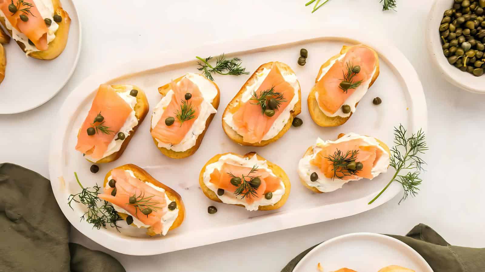 A platter of smoked salmon bruschetta topped with cream cheese, capers, and dill, surrounded by capers and herbs on a white background.