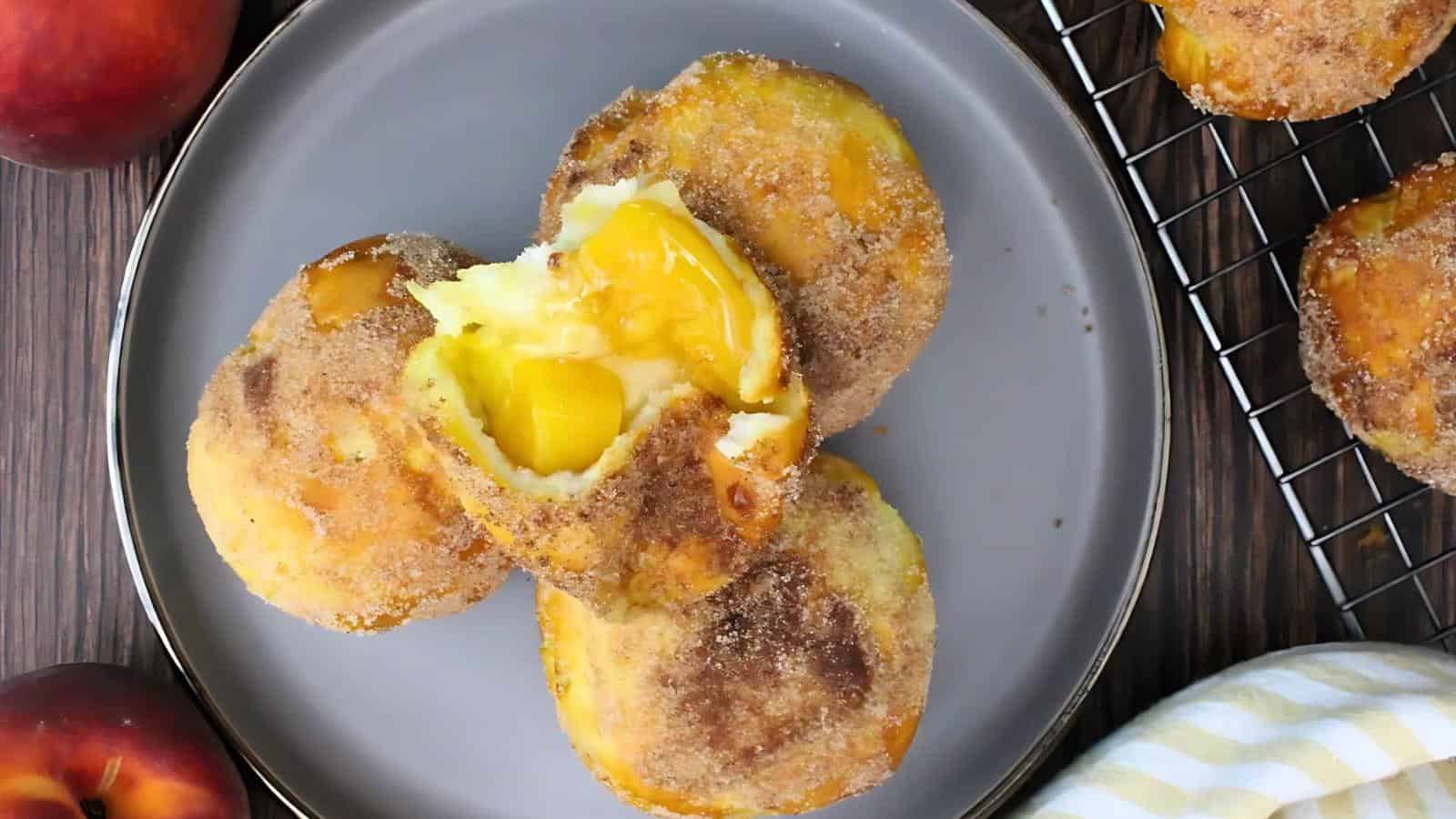 Peach-filled pastries coated with cinnamon sugar are stacked on a gray plate, with a cooling rack and peaches nearby.