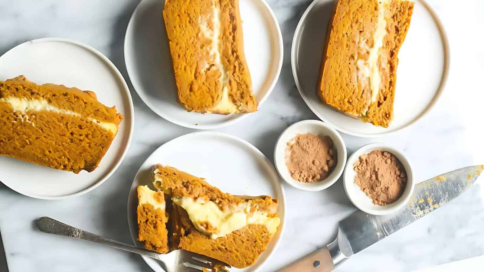 Slices of pumpkin roll cake with cream cheese filling on plates, accompanied by small bowls of cocoa powder.