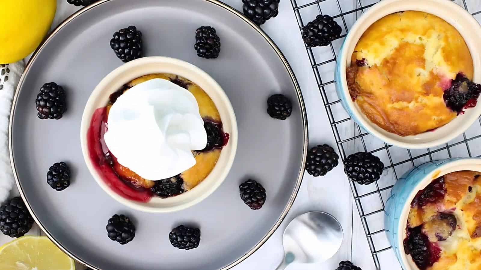 A plate with a small lemon blackberry clafoutis topped with whipped cream, surrounded by fresh blackberries. Two more clafoutis on a cooling rack are nearby.