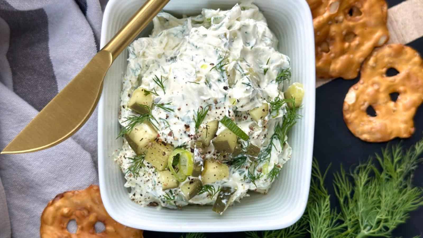 Bowl of creamy salad with chopped dill and cucumbers, surrounded by pretzel crisps, on a dark surface with a striped cloth and a gold knife.