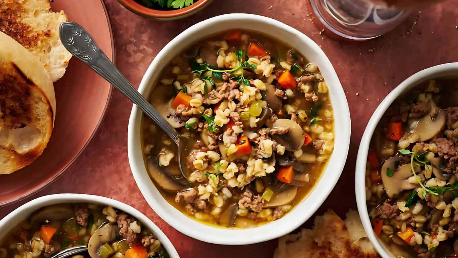 A bowl of vegetable and barley soup on a table with a spoon, surrounded by slices of toasted bread and two small bowls of soup.
