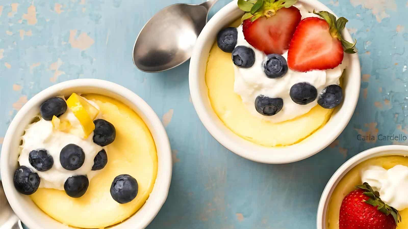 Three bowls of cheesecake topped with whipped cream, blueberries, and strawberries on a light blue surface, accompanied by a silver spoon.