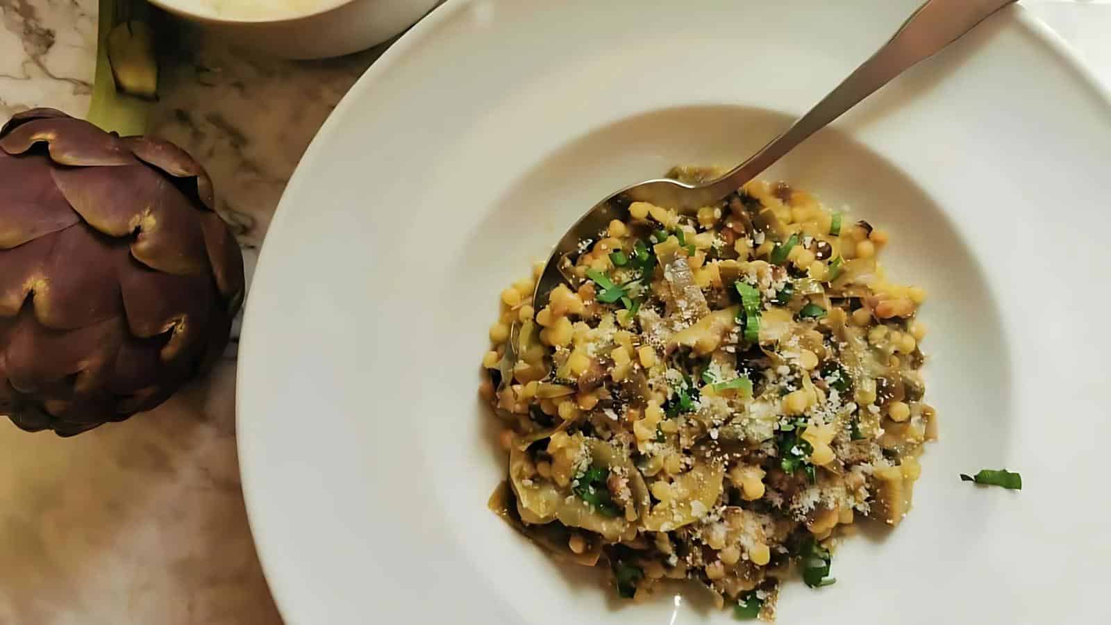 A plate of pasta with lentils and chopped herbs, topped with grated cheese. An artichoke and a bowl of grated cheese are nearby on the table.