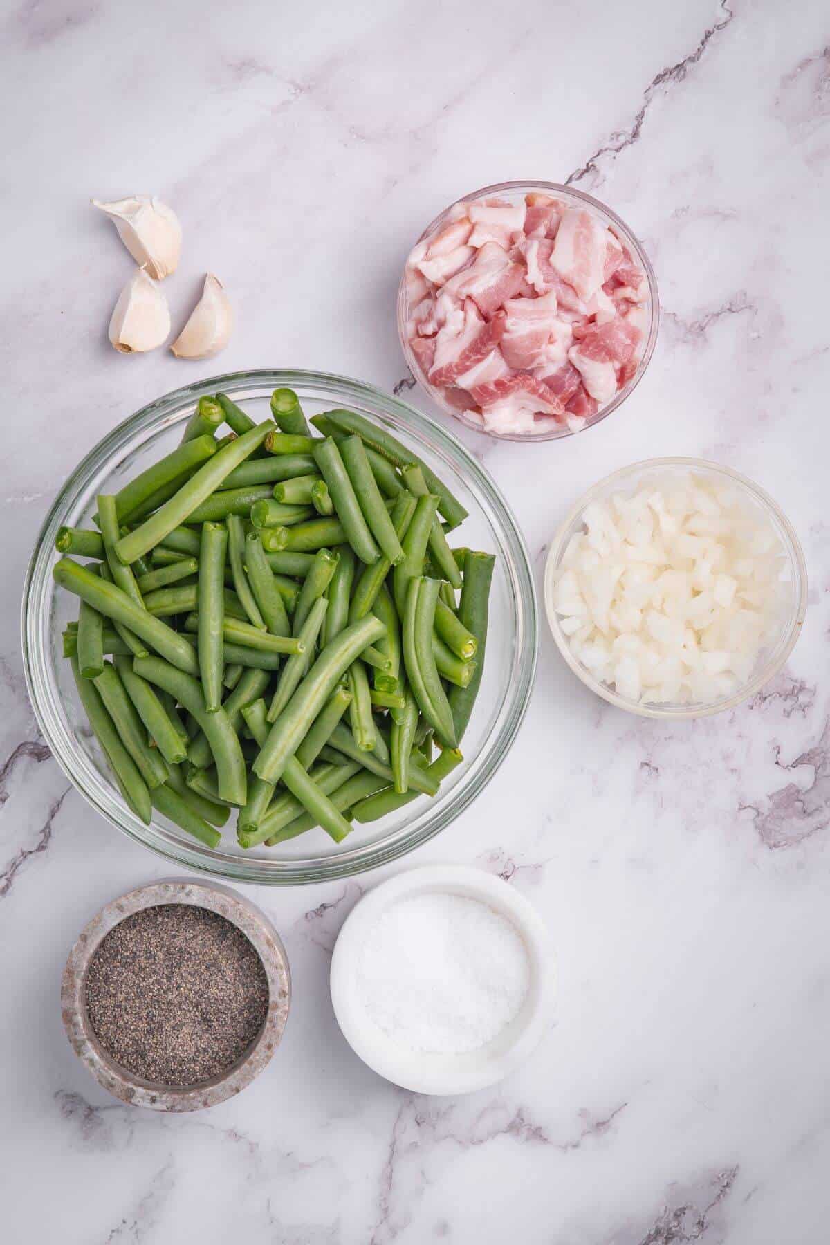 Ingredients on a marble surface: fresh green beans in a bowl, diced onions, chopped bacon, garlic cloves, and small bowls of salt and pepper.