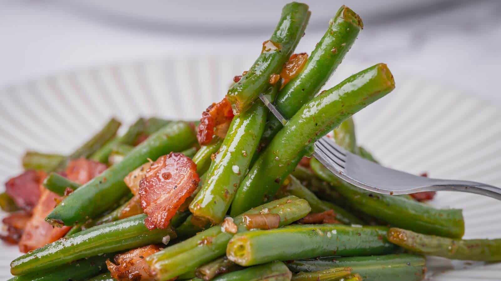 A fork lifts green beans and bacon pieces from a white plate.