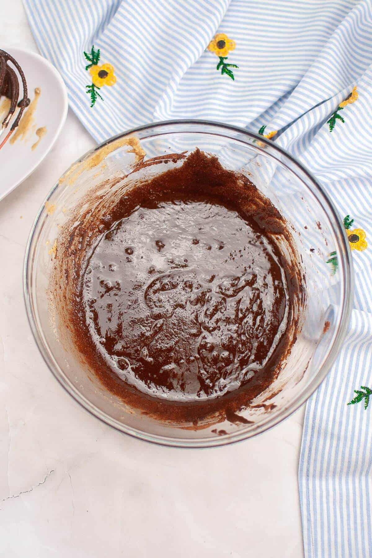 A glass bowl filled with smooth chocolate batter on a table with a blue and white striped cloth, adorned with yellow flower patterns.