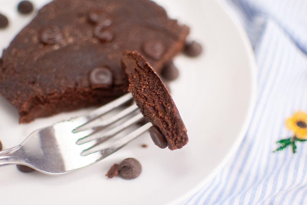 A fork holding a bite of chocolate brownie on a white plate, surrounded by chocolate chips. A blue striped cloth with a small embroidered flower is partially visible.