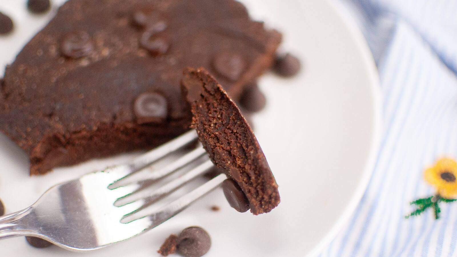 Fork holding a slice of a chocolate brownie with chocolate chips on a white plate.