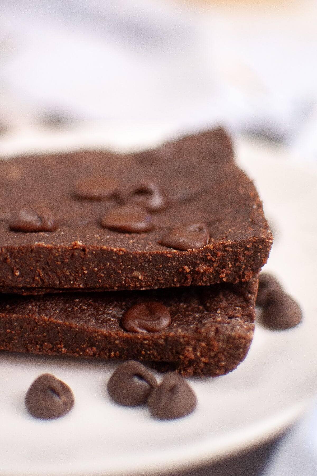 Two pieces of chocolate brownie stacked on a plate, topped with chocolate chips. Additional chips are scattered around.