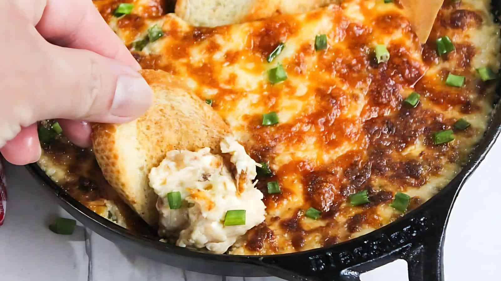 Close-up of a cheesy baked dip topped with chopped green onions, with a hand dipping a piece of toasted bread into it, in a black skillet.