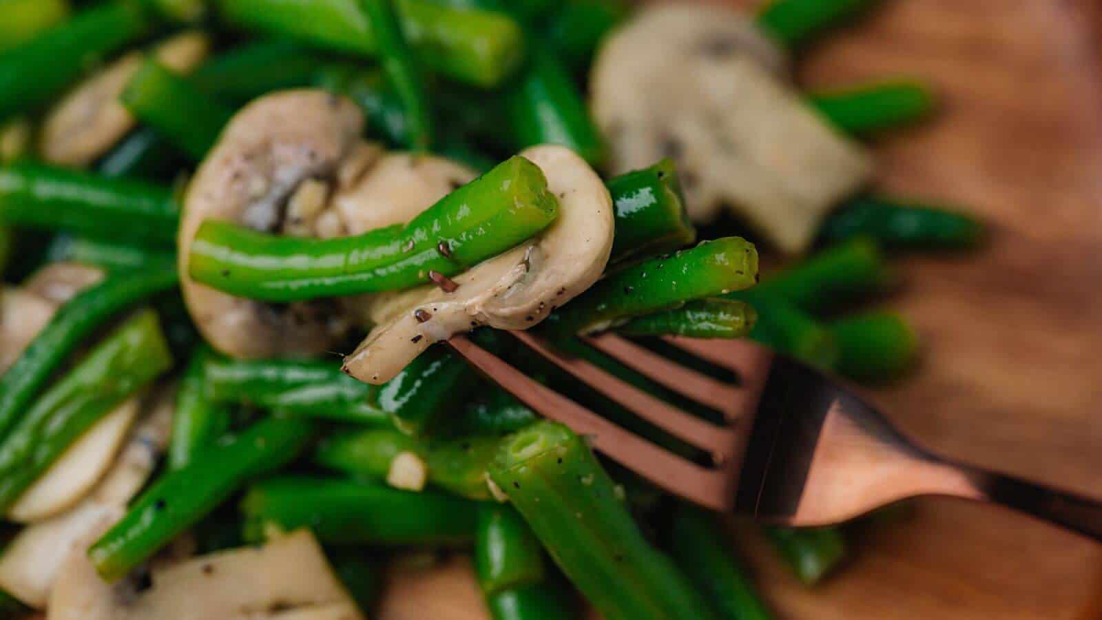 A fork holds cooked green beans and sliced mushrooms, with more on a plate in the background.