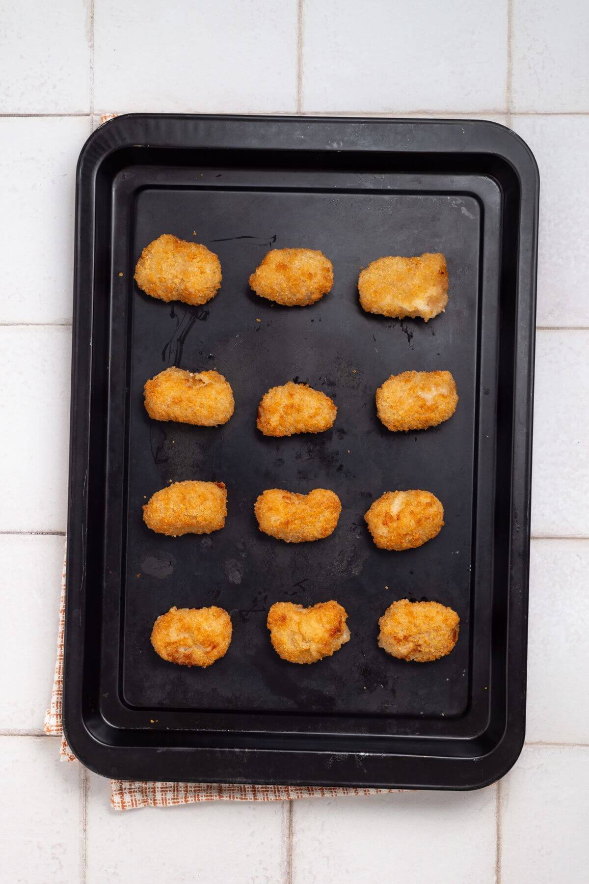 A black baking tray with twelve evenly spaced, golden-brown, breaded chicken nuggets on a tiled surface.
