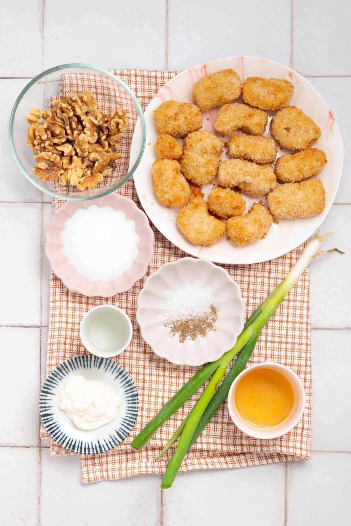 Ingredients arranged on a checkered cloth, including breaded nuggets, walnuts, salt, pepper, mayonnaise, green onions, and a small bowl of orange sauce.