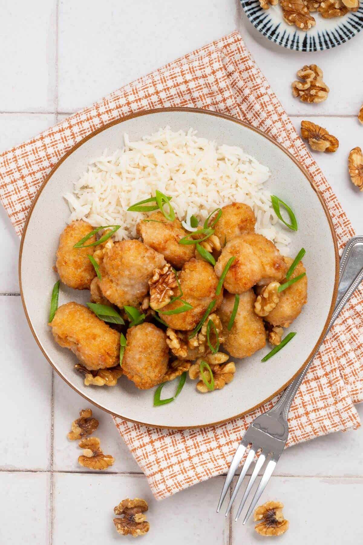 Plate of breaded chicken pieces with walnuts, sliced green onions, and white rice on a checkered napkin, accompanied by a fork.