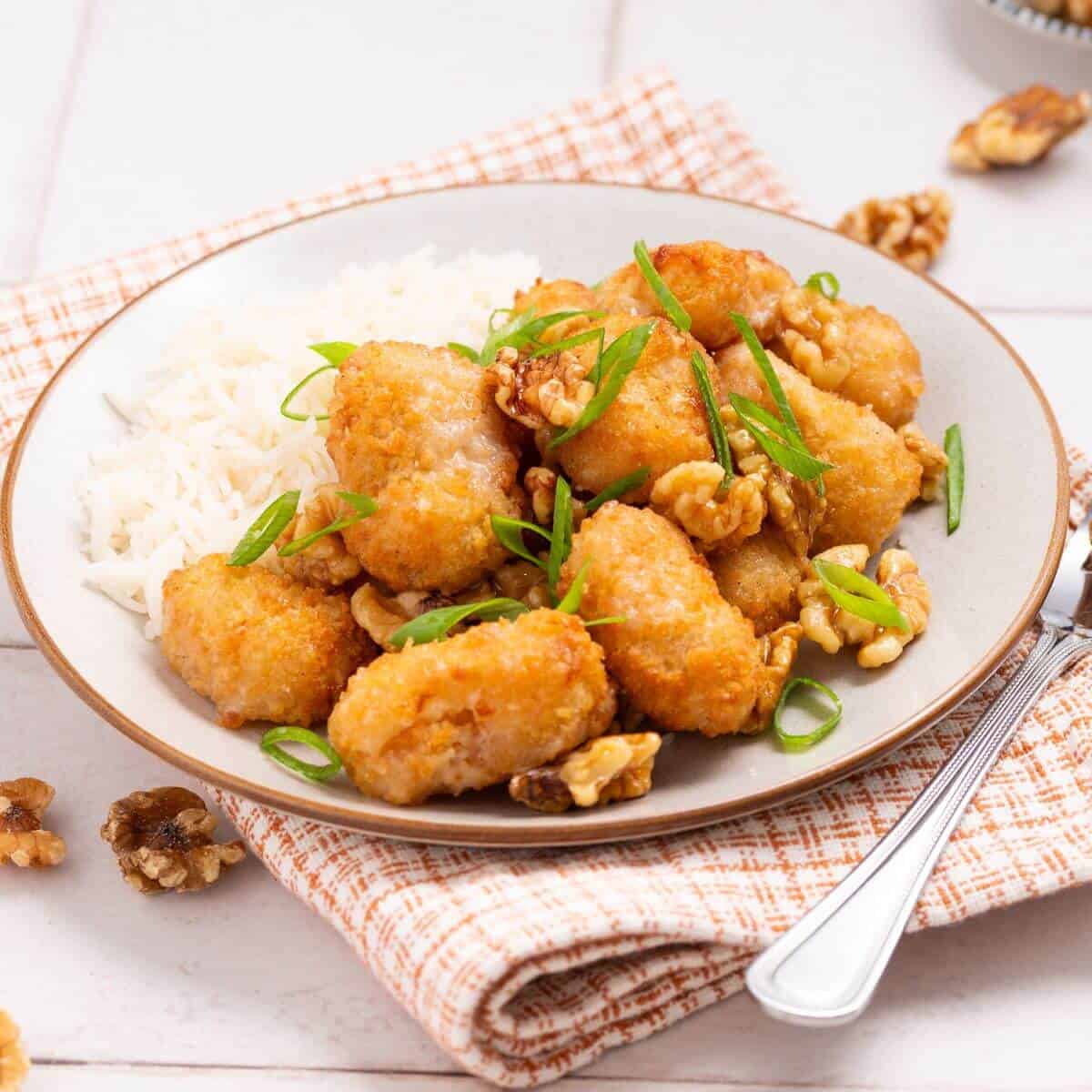 A plate of fried chicken pieces with walnuts and green onions, served with white rice on a patterned cloth.