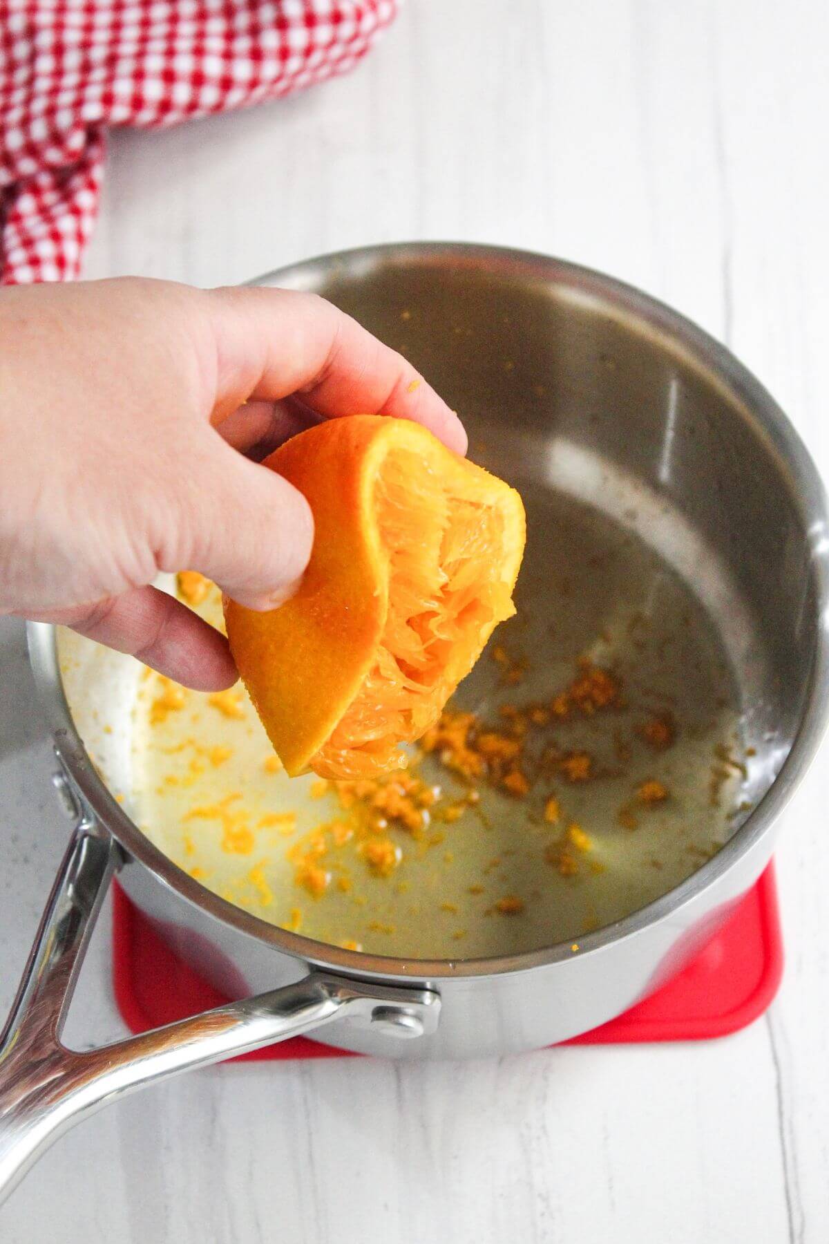 Hand squeezing an orange half over a metal saucepan with orange zest inside, placed on a red trivet.