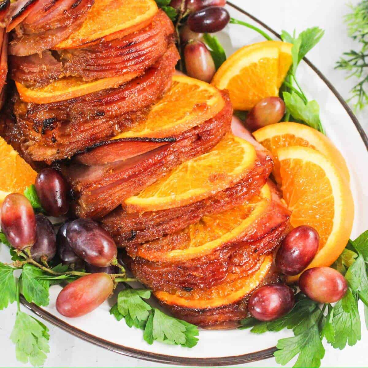 Sliced ham garnished with orange slices, red grapes, and parsley on a white platter.