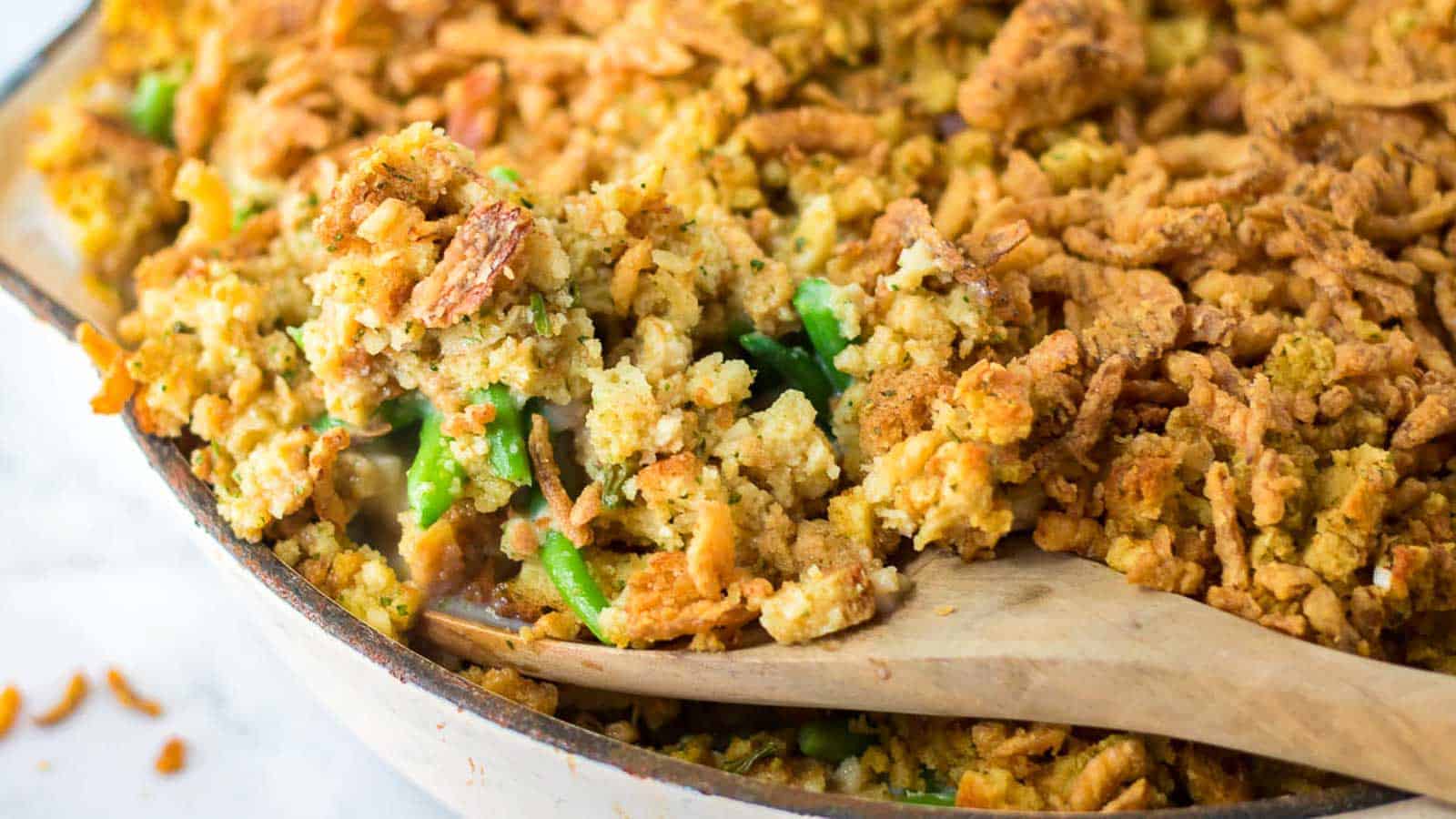 A wooden spoon scoops a serving of green bean casserole topped with crispy breadcrumbs from a baking dish.