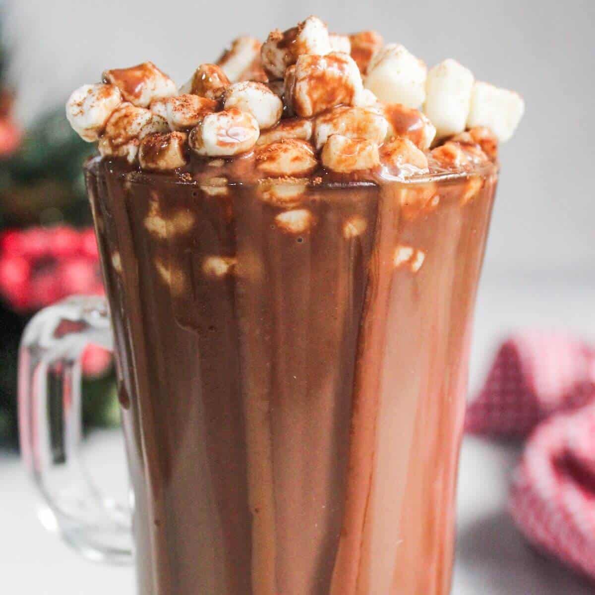 A glass mug filled with hot chocolate, topped with mini marshmallows and drizzled with chocolate syrup.
