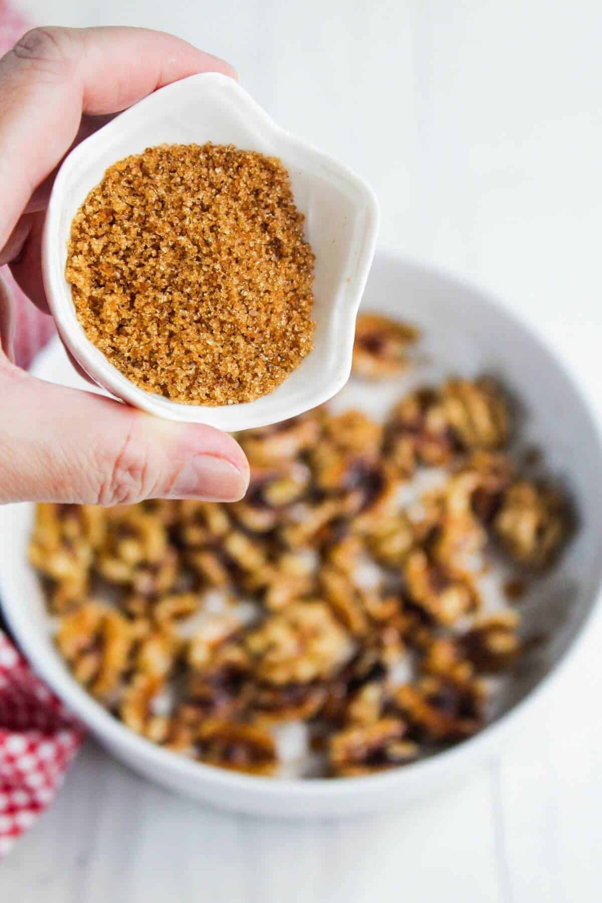 A hand holds a small white bowl filled with brown sugar above a dish of walnuts.