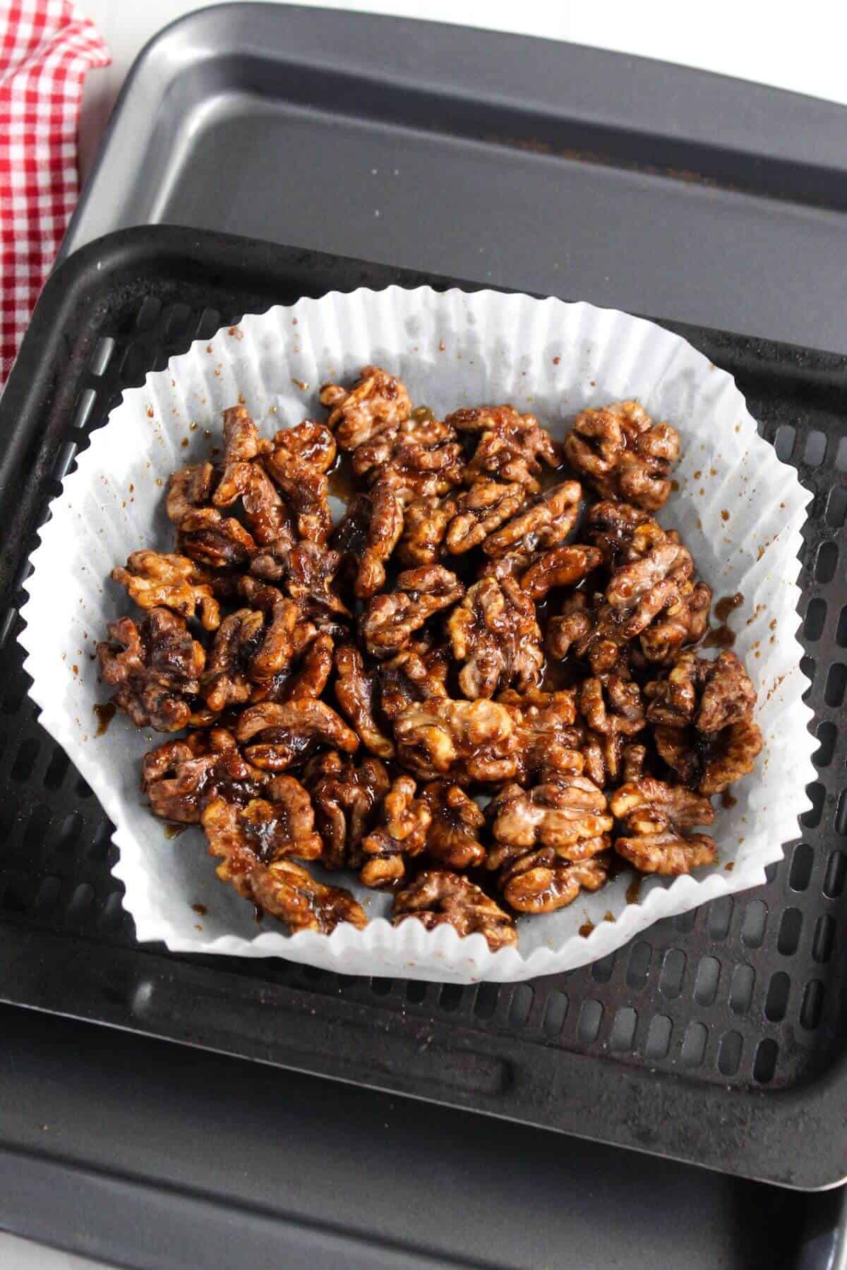 A bowl of glazed walnuts sits on a baking tray lined with white parchment paper. A red and white checkered cloth is partially visible in the corner.