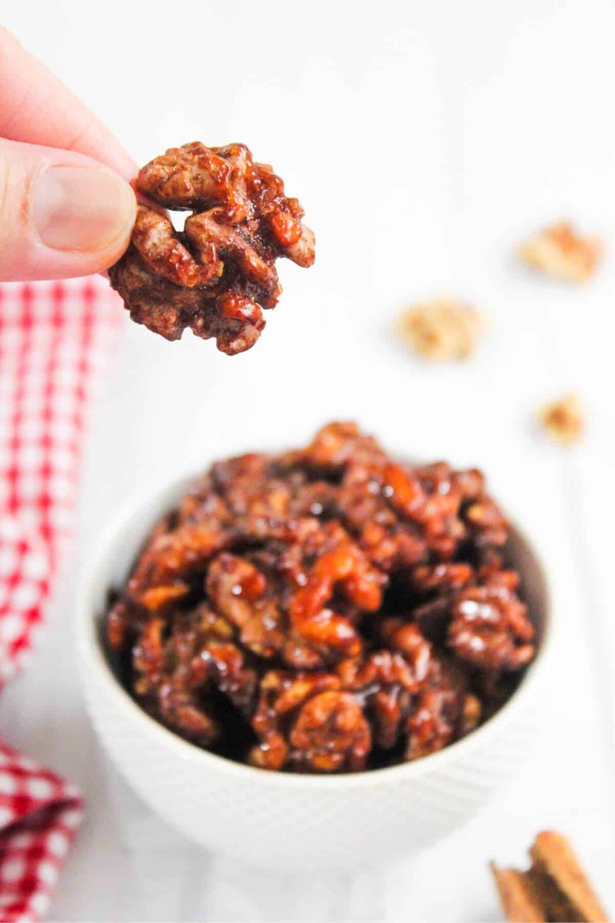 A hand holds a piece of candied walnut above a bowl of more candied walnuts on a white surface with a red checkered cloth nearby.