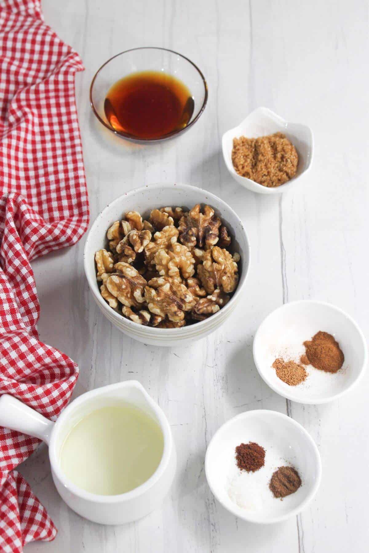 Ingredients on a table: walnuts in a bowl, syrup in a glass dish, brown sugar in a small bowl, spices in two small bowls, and a jug of milk, next to a red checkered cloth.
