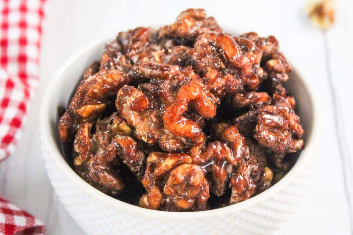 A bowl of glazed roasted nuts on a white surface, with a red checkered cloth partially visible on the left.