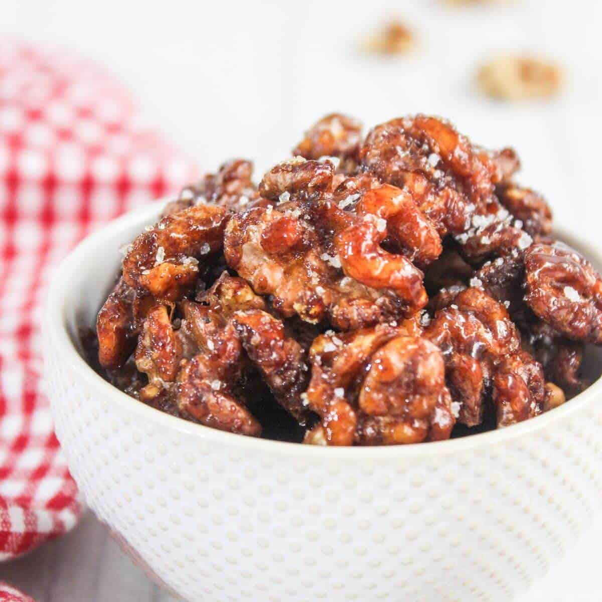 A white bowl filled with glazed candied walnuts, with a red and white checkered cloth beside it.