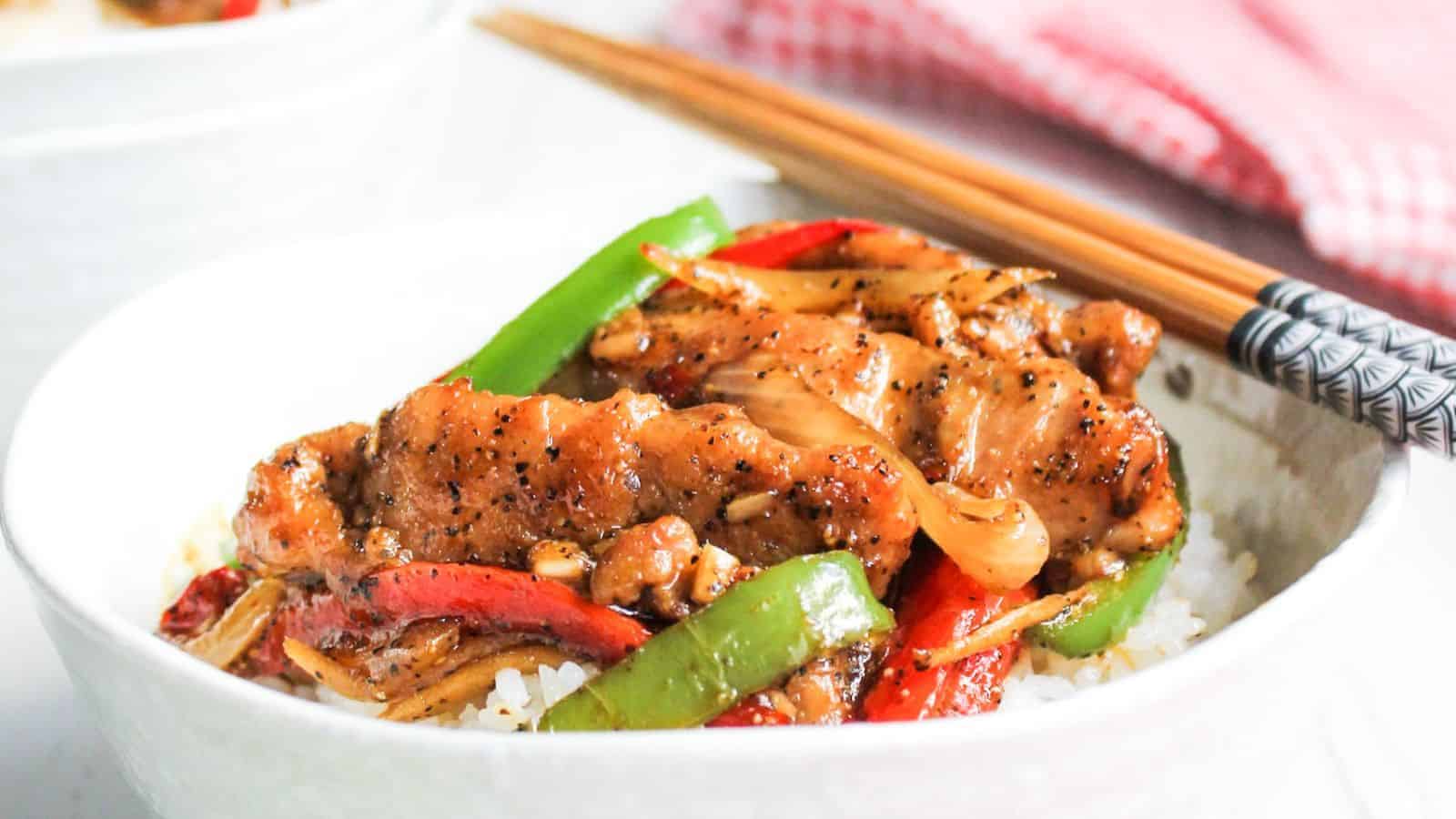 A bowl of stir-fried chicken with green and red bell peppers on white rice, with chopsticks on the side.