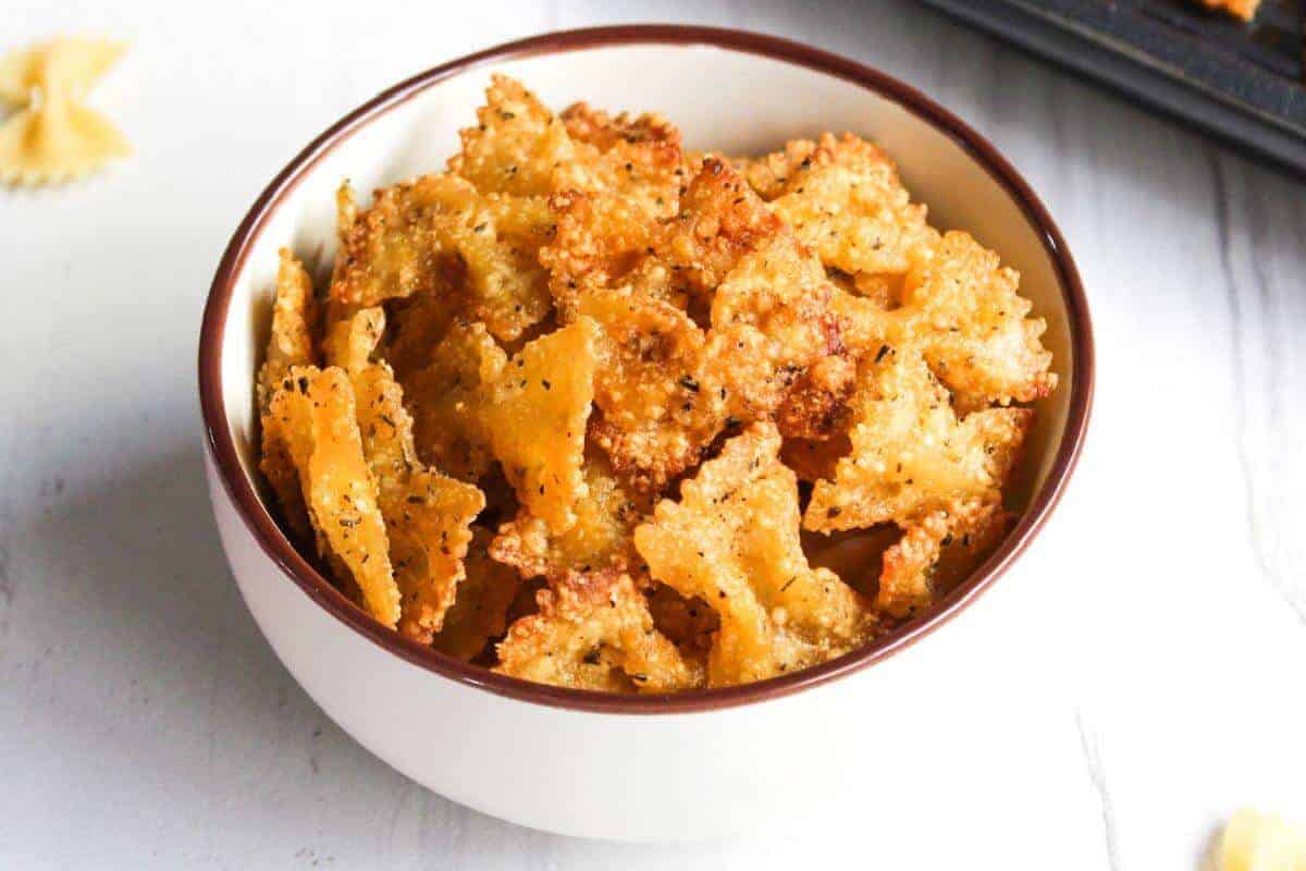 A bowl of crispy seasoned bow-tie pasta chips on a light surface.