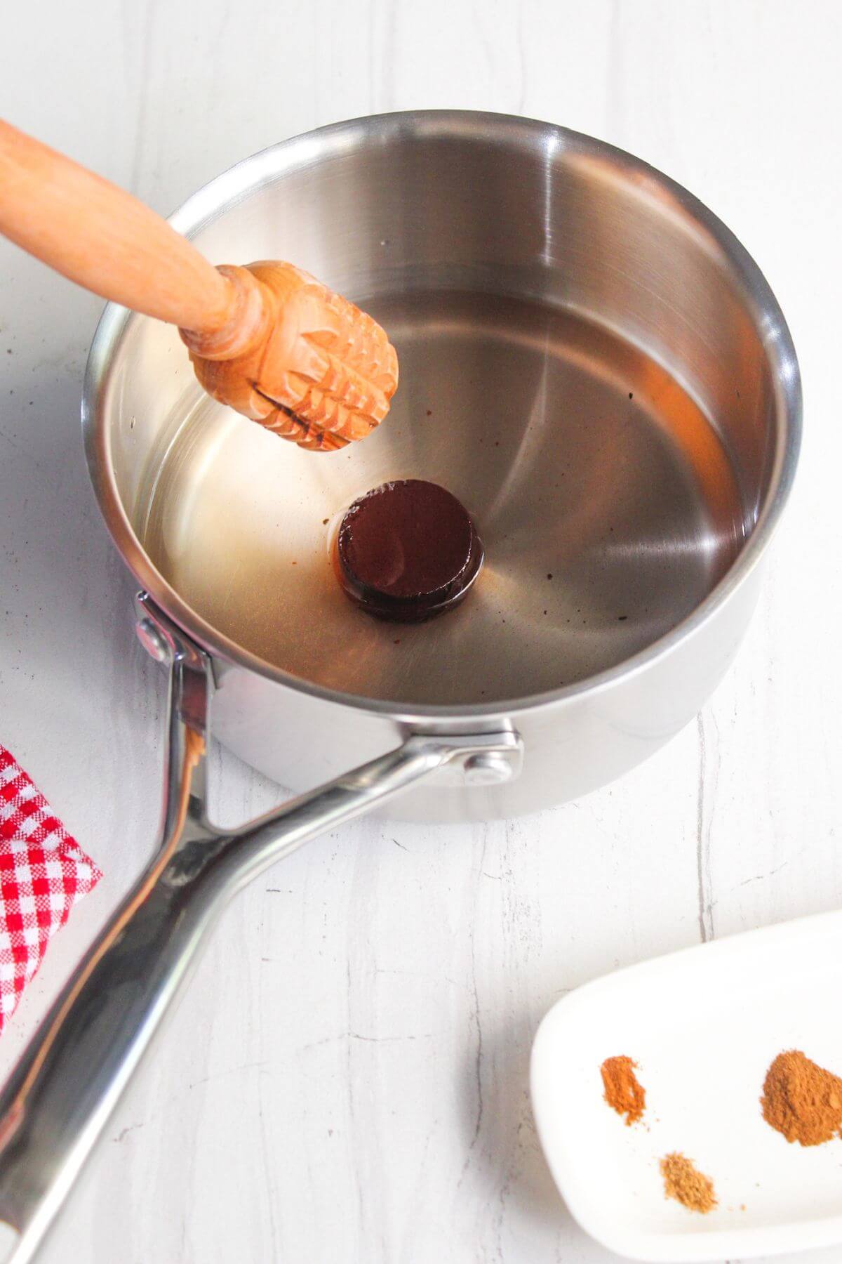 A round chocolate piece in a saucepan of water with a wooden tool, next to spices on a white dish.