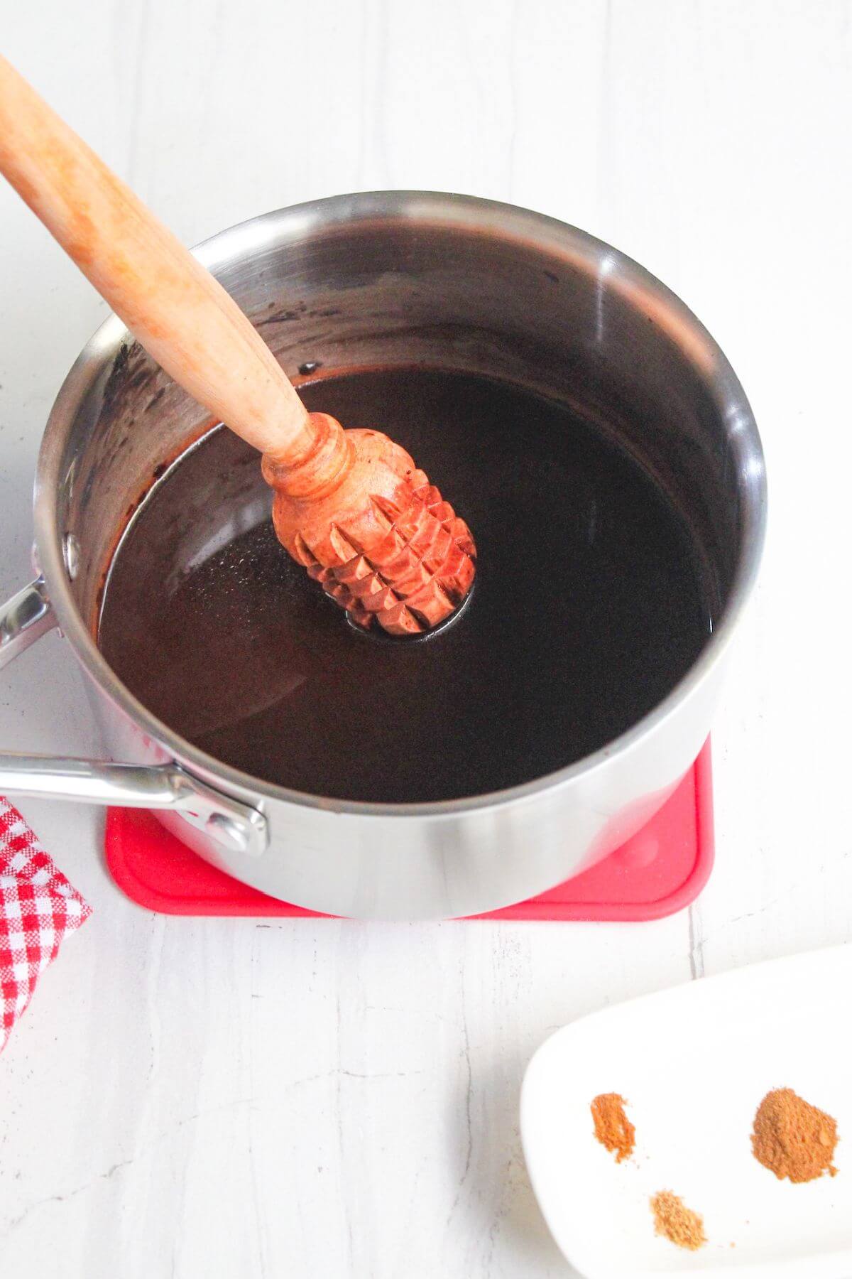 A pot of dark liquid with a wooden whisk on a red trivet. A plate with spices is nearby on a white surface.
