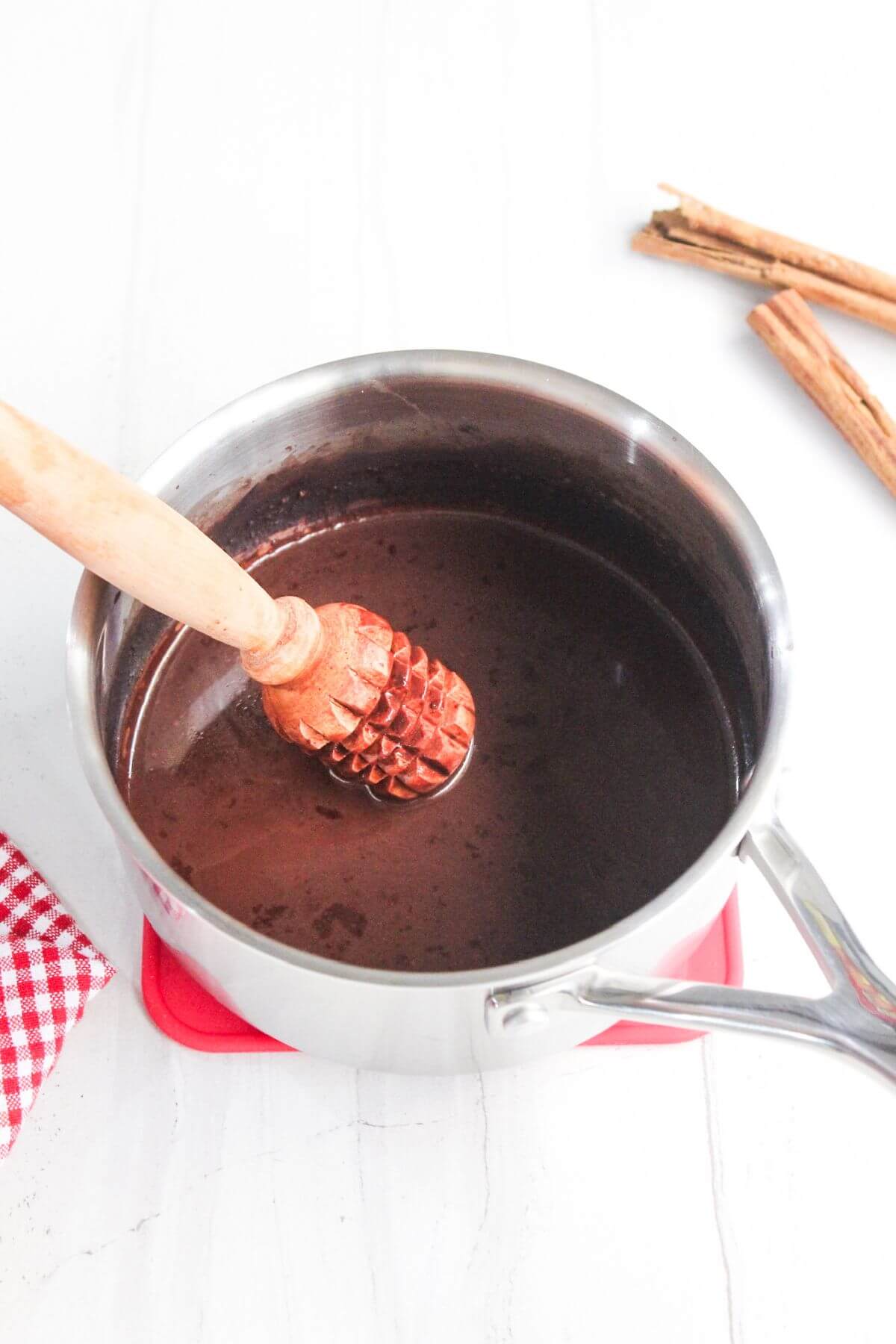 A pot of chocolate sauce with a wooden whisk sits on a red trivet. Cinnamon sticks are in the background, and a red checkered cloth is visible.