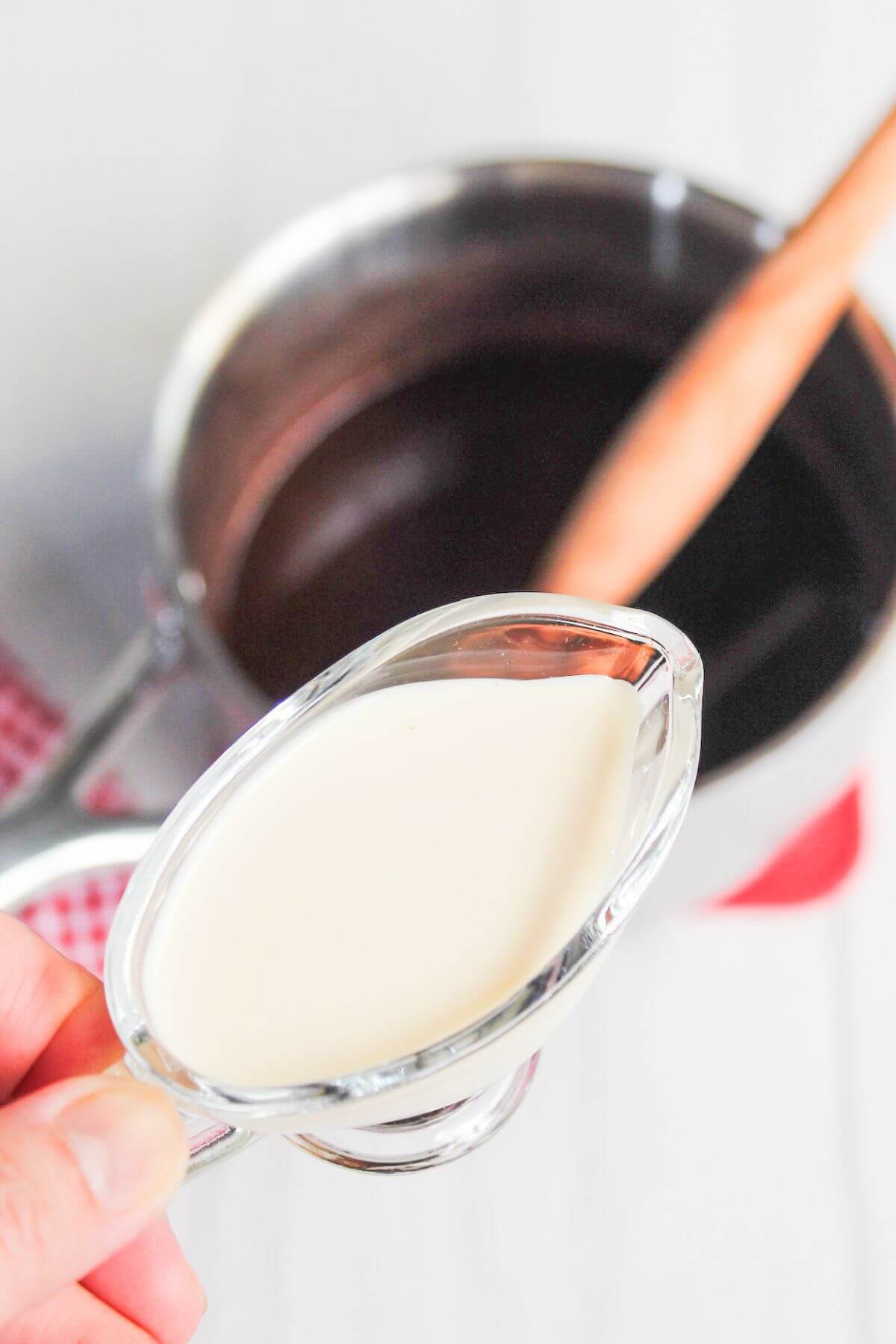 A small glass pitcher of cream is being poured into a pot of chocolate with a wooden spoon in the background.