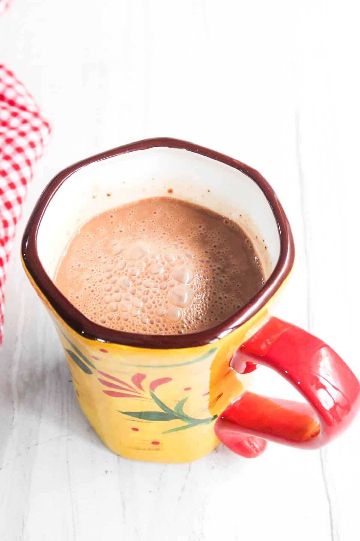A hexagonal ceramic mug with a yellow floral design and red handle is filled with frothy hot chocolate, placed on a white surface next to a red and white checkered cloth.