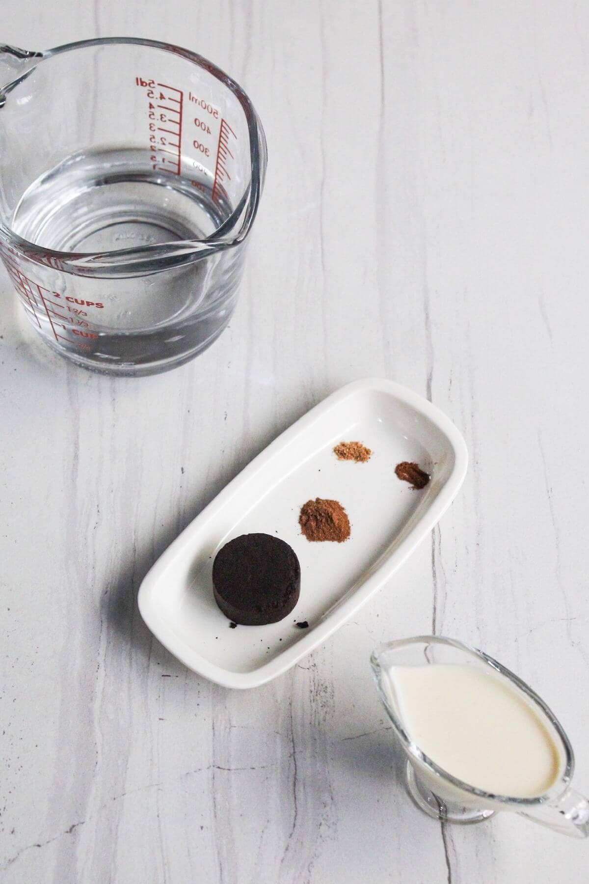 Measuring cup with water, a white dish with a chocolate piece and spices, and a small glass pitcher of milk on a light surface.