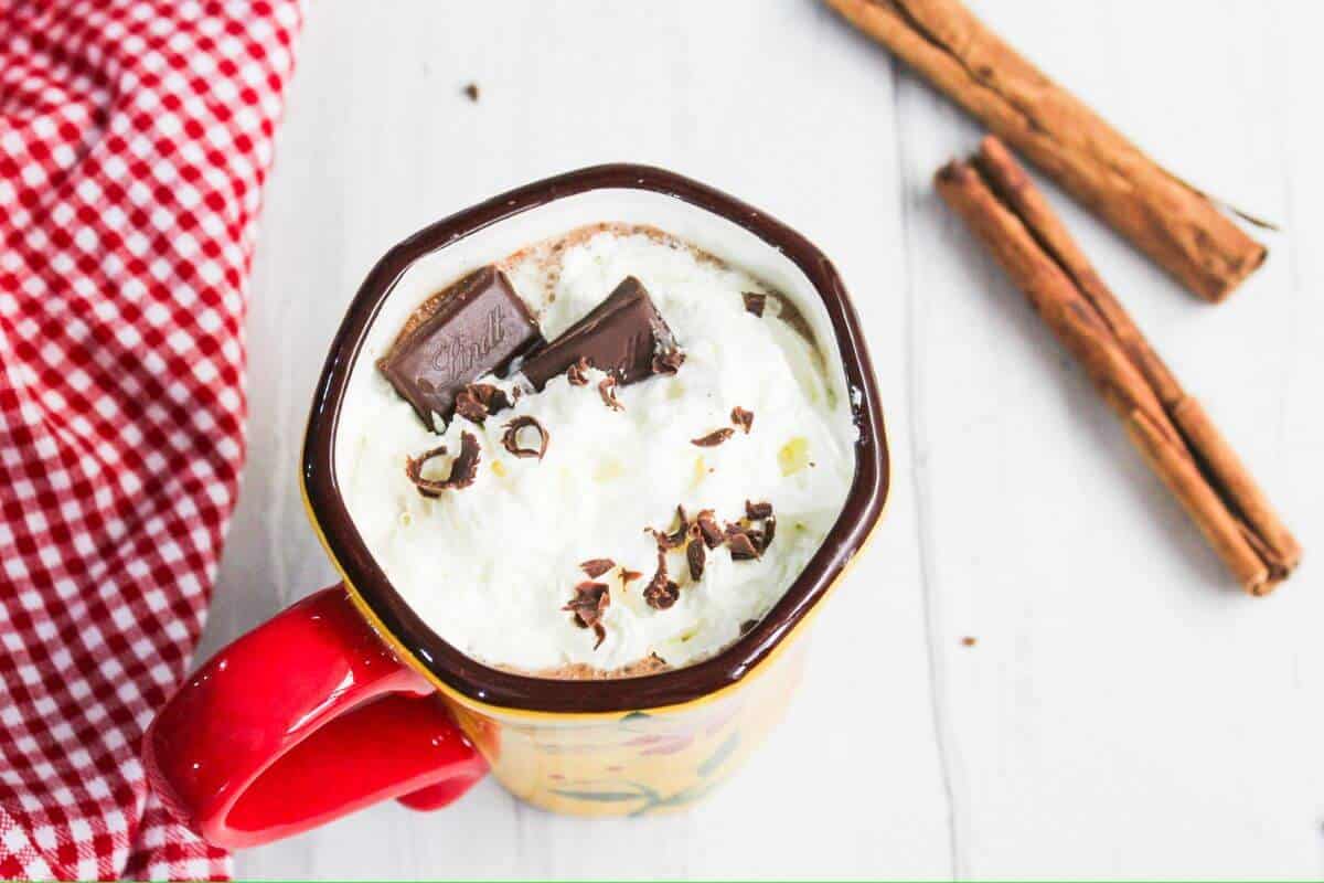 A mug of hot chocolate with whipped cream and chocolate pieces, next to cinnamon sticks on a white surface with a red checkered fabric.